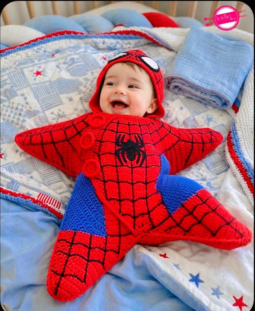A baby wearing a red and blue crocheted star-shaped Spider-Man costume with a matching hood, lying on a blue patterned blanket.